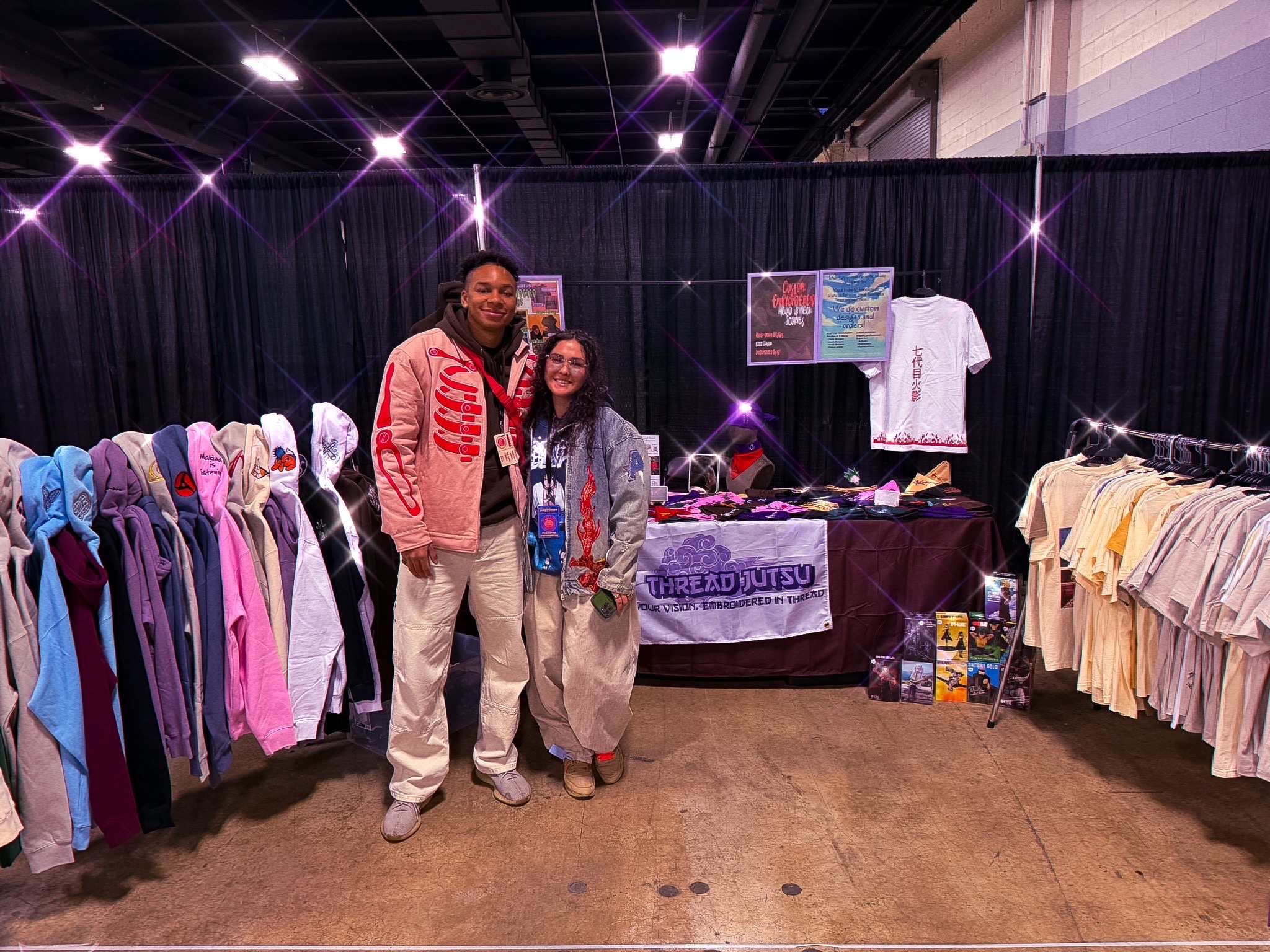 The two creators of Thread Jutsu at their booth, surrounded by embroidered hoodies and tees